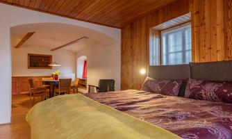 A cozy bedroom with wood paneling and a large bed. In the background, there is a dining area with chairs and a table.