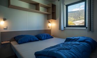 A cozy bedroom with a double bed and blue cushions. Through the window, there is a beautiful view of the mountains.
