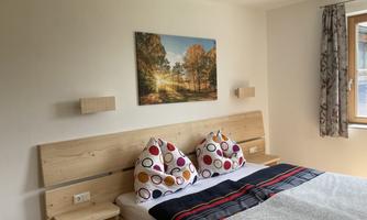 A cozy bedroom with a double bed, colorful pillows, and a wooden frame. A picture of an autumn landscape hangs on the wall.