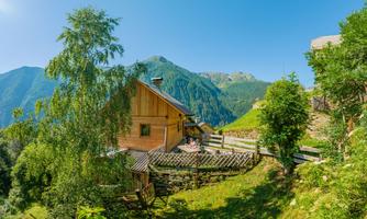 A rustic wooden house surrounded by green mountains and trees. The sun is shining and the sky is clear and blue.