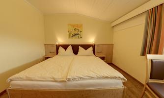 A cozy bedroom with a large double bed and a bright, friendly ambiance. A colorful picture hangs on the wall, and the windows are adorned with colorful curtains.