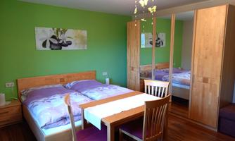 A cozy bedroom with a double bed, table, and chairs. The walls are painted green and there is a large wardrobe.