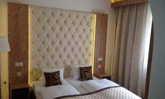 A modern bedroom with a padded headboard and warm light. The two beds are adorned with elegant pillows, and curtains let in soft light.