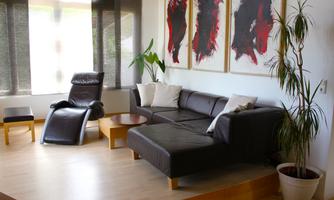 A modern living space with a black couch and a comfortable armchair. Large windows provide plenty of natural light, and interesting artworks adorn the walls.