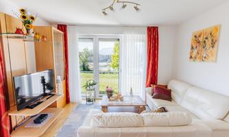 A modern living room with a white sofa and a television. Large windows provide a view of the green landscape outside.