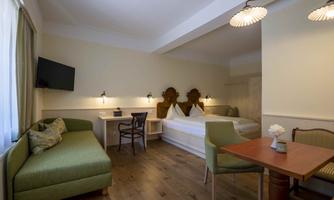 A bright room with a double bed, a desk, and a cozy sofa. The room is modernly furnished and has large windows that let in a lot of light.