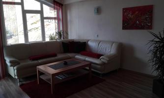 A modern living room with a beige couch and red pillows. A wooden table sits in the center, surrounded by large windows and plants.