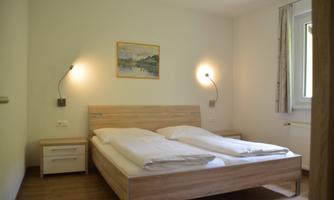 A simple bedroom with a double bed and a bedside table. The walls are light, and there is a landscape painting on the wall.