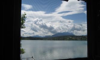 A view through a window of a calm lake under a clear sky with clouds. In the background, gentle mountains are visible.