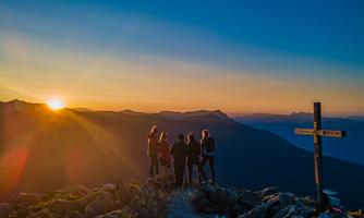 A group of people stands on a mountain peak and watches the sunset. In the background, mountains and a cross are visible.