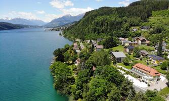 A picturesque landscape with a clear lake and green shores. On the shores, there are houses and trees, surrounded by mountains.