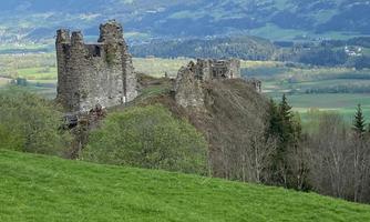 An old castle ruin on a hill, surrounded by green meadows. In the background are majestic mountains and a clear sky.