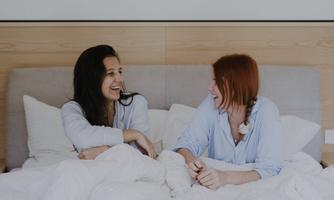 Two women are sitting laughing in bed under a blanket. They are wearing comfortable nightwear and enjoying a cozy time together.