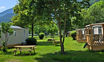 A quiet campsite with green meadows and shade-giving trees. In the background, mobile homes and high mountains can be seen.