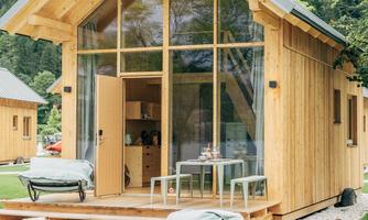 A modern wooden house with large windows and a green outdoor area. There are loungers on the lawn, surrounded by trees.