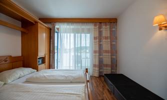A bright bedroom with two single beds and a large window. The furnishings are made of wood, and there is a small seating area next to the window front.