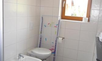 A modern bathroom with white tiles and a window. There is a toilet and a bidet, as well as a drying rack.
