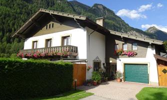 A charming house in alpine style, surrounded by green mountains. The balcony is adorned with flowers, and a garage door is visible.