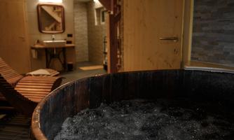 A relaxing wellness area with a wooden bathtub and bubbling water. In the background, there is a lounger and a modernly designed bathroom.