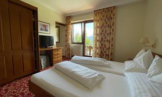 A cozy hotel room with two single beds and a television. A large window with curtains provides daylight and a view outside.