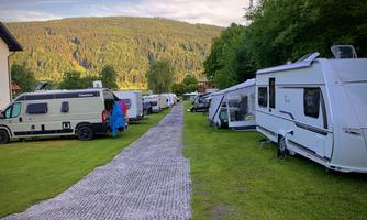 A campsite with several motorhomes and caravans along a well-maintained path. In the background, green hills and trees can be seen.
