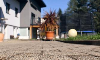 A paved terrace in front of a modern building. In the foreground, there is a flower pot and a large ball, while in the background, a trampoline can be seen.