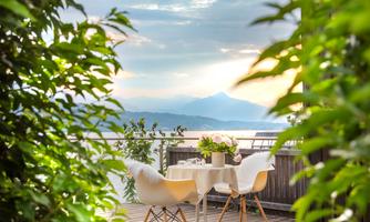 An inviting terrace with chairs and a small table, surrounded by green plants. The view of the landscape is peaceful and the sky is slightly cloudy.