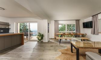 A modern living room with an open kitchen and large windows. The room is bright and provides access to a balcony with a view of greenery.