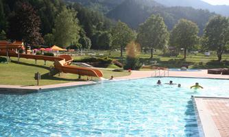 A beautiful swimming pool with slides and green meadows in the background. The surroundings are framed by mountains and trees.