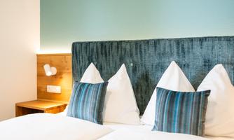 A cozy room with two beds and cushions in white and blue. An open book lies on the bed, while the wall is painted in a soft blue.