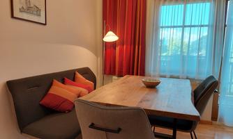 A cozy dining area with a wooden table, modern chairs, and a couch. Large windows provide plenty of daylight and an inviting atmosphere.