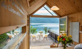 A cozy wooden house with large windows and a view of a lake. Outside, there is a table and chairs, surrounded by flowers and nature.