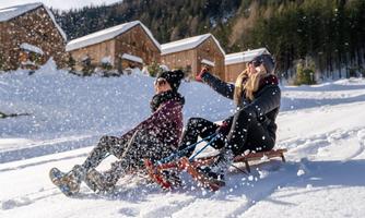 Two women are happily sledding in the snow and enjoying the winter sun. In the background, there are wooden chalets and snow-covered trees.