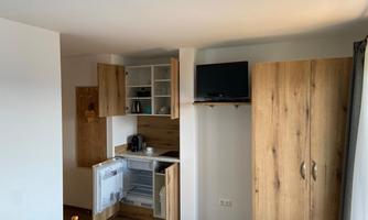 A modern kitchen nook with wooden cabinets and a small refrigerator. A television is mounted on the wall.
