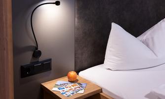A cozy bedroom with a pillow and a bedside table. On the table are postcards and an apple, next to a reading lamp and power outlets.