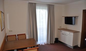 A bright room with a table, chairs, and a television on the wall. Large windows with curtains provide plenty of daylight.