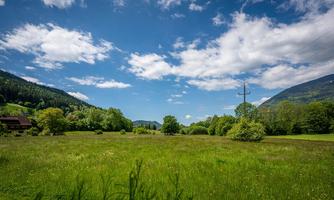 A vast green field under a clear blue sky. In the background, gentle hills and trees can be seen.