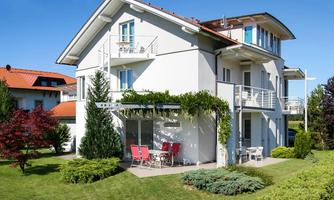 A modern two-story house with a garden. In front of the terrace, there are red chairs and a table.