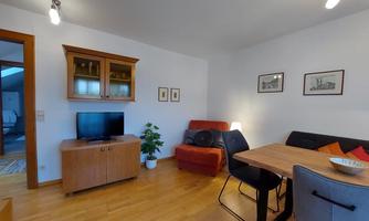 A cozy living room with wooden flooring and modern furniture. A television is placed next to a large window, and a dining table with chairs is located in the center of the room.