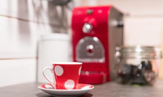 A red coffee cup with white dots is standing on a saucer. In the background, a red coffee machine can be seen.