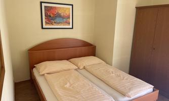 A simple bedroom with a double bed and two pillows. A colorful picture hangs on the wall, and a wardrobe stands in the background.