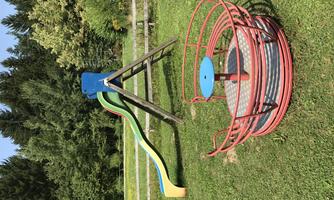A playground with a colorful slide and a spinning plate. Surrounded by green grass and trees.