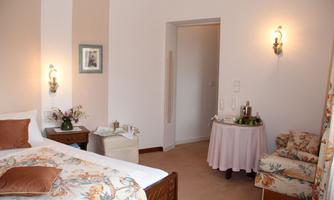 A cozy bedroom with a large bed and floral details. In the background, there is a seating area and a small table with refreshments.