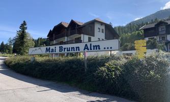 A sign with the inscription "Mai Brunn Alm" stands by a road. In the background, several houses and a green landscape can be seen.