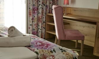 A cozy room with a bed and an interesting floral pattern on the bedspread. Next to it is a desk with a pink chair and a lamp.