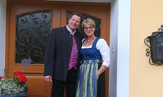 A couple stands in front of a front door wearing traditional clothing. The woman is wearing a dirndl and the man is wearing a suit with a vest.