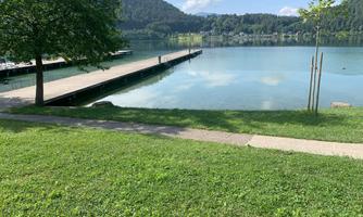 A calm lake with a wooden pier and gentle hills in the background. The water is clear and reflects the surroundings.