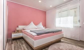 A bright bedroom with pink walls and a large bed. The decor is modern and simple, with wooden flooring and beautiful curtains.