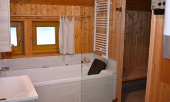 A modern bathroom with wooden paneling and tiles. The bathtub and sink are well-equipped, and a sauna is visible in the background.