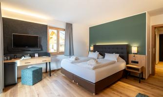A modern hotel room with a large bed, a desk, and a television. The decor is stylish and bright, with wooden flooring and neutral colors.